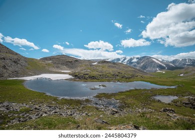 Beautiful small lake with snowfield among green grassy hills and rocks with view to snow-capped mountain range. Scenic landscape with alpine lake and snowy high mountains. Mountain lake in highlands. - Powered by Shutterstock