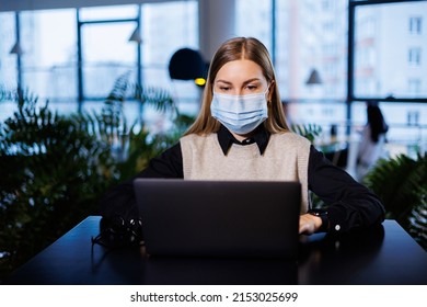 A Beautiful Smart Businesswoman In A Large Company Holds An Online Meeting With Business Partners In A Protective Mask During The Coronavirus, She Sits At A Table At A Workplace With A Laptop