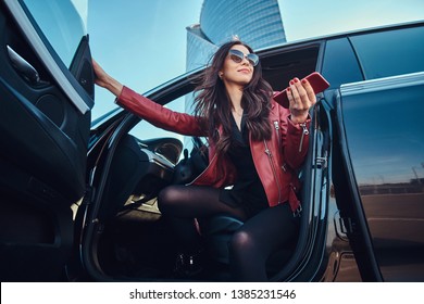 Beautiful smart women is posing in her new car while chatting on mobile phone. She is wearing red leather jacket and sunglasses.