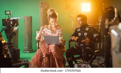 Beautiful Smiling Actress Wearing Renaissance Dress And Actor Wearing Motion Capture Suit Having Lunch Break, Sitting On Chairs, Use Laptop And Talk. Studio High Budget Movie. Costume Drama Film Set