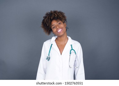 Beautiful Smiling African American Doctor Female With Broad Smile, Shows White Teeth, Wears Medical Uniform, Stands Over Gray Studio Wall Rejoices Having Day Off.