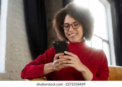 Beautiful smiling African American woman holding mobile phone watching videos sitting at home. Modern female wearing red eyeglasses using mobile app, shopping online, ordering food. Mobile banking - Powered by Shutterstock
