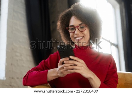 Beautiful smiling African American woman holding mobile phone watching videos sitting at home. Modern female wearing red eyeglasses using mobile app, shopping online, ordering food. Mobile banking