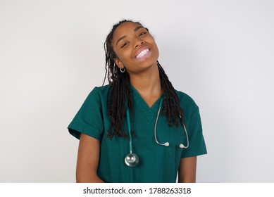 Beautiful Smiling Afro American Doctor Female With Broad Smile, Shows White Teeth, Wears Medical Uniform, Stands Over White Studio Wall Rejoices Having Day Off.