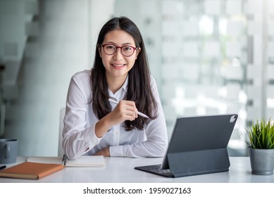 Beautiful smiling Asian businesswoman wearing  glasses hand holding pen working using tablet at office. Looking at camera.