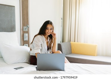 Beautiful Smiling Asian Girl With Long Hair Working On Laptop On Bed In Hotel Room