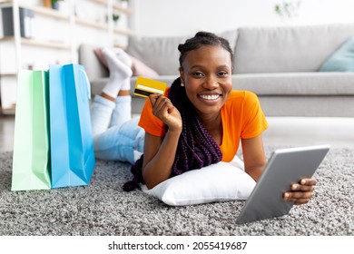 Beautiful Smiling Black Lady Lying On Floor With Digital Tablet, Credit Card And Shopping Bags At Home. Cheery Young Woman Purchasing Goods Online In Web Store, Making Remote Payment