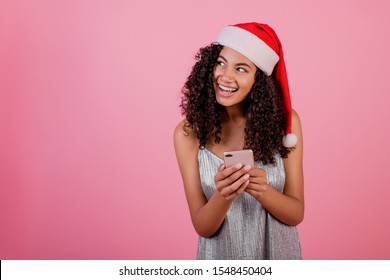 beautiful smiling black woman with phone wearing santa hat and dress isolated over pink - Powered by Shutterstock