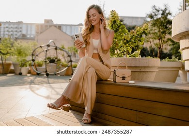 beautiful smiling blond woman sitting in sunny city street with phone, urban fashion style, summer trend season, business lady, glam hair hairstyle, beige suit wearing vest and trousers - Powered by Shutterstock