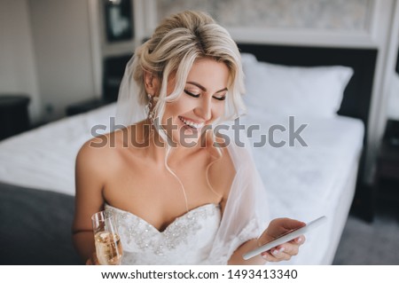 A beautiful, smiling blonde bride in a white dress is sitting on the bed and looking at the phone, reading a message and drinking champagne. Wedding portrait of a cheerful girl.