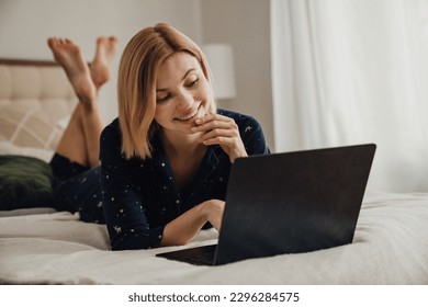 Beautiful smiling blonde woman dressed in pajamas working on laptop while laying in bed in her apartment - Powered by Shutterstock