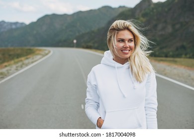 Beautiful smiling blonde young woman traveler in white hoodie on road, trip to mountains, Altai