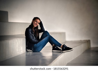 Beautiful smiling brunette model girl in stylish black leather jacket and dark blue jeans sitting on the concrete stairs with large steps. - Powered by Shutterstock