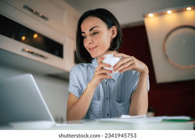 Beautiful smiling brunette woman with cup in hands drinks coffee or tea while watching film on the laptop at home on the kitchen - Powered by Shutterstock