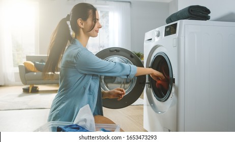 Beautiful Smiling Brunette Young Woman Sits In Front Of A Washing Machine In Homely Jeans Clothes. She Loads The Washer With Dirty Laundry. Bright And Spacious Living Room With Modern Interior.