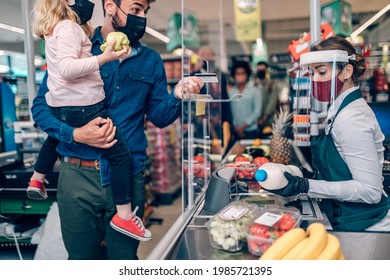 Beautiful Smiling Cashier Working At A Grocery Store. Father Is Holding His Daughter At Market's Checkout While Paying By Debit Card. They Are All Wearing Face Protective Masks.