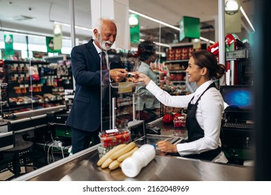 Beautiful Smiling Cashier Working At A Grocery Store. Senior Man At Market's Checkout Paying By Debit Card.