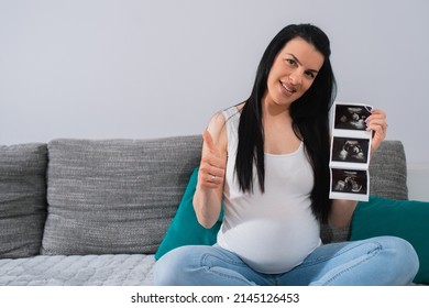 Beautiful Smiling Caucasian Pregnant Woman Shows OK To Camera With The Thumb On Her Right Hand And Holds And Shows Ultrasound Pictures Of Her Unborn Baby With Her Left Hand. She Sits With Crossed Legs