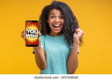 Beautiful Smiling Dark Skinned Woman Showing Sincere Excitement About Victory At Bookmaker's Website. Girl Being Happy Winning Bet In Online Sport Gambling Application On Her Mobile Phone.