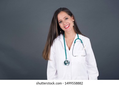 Beautiful Smiling Doctor Female With Broad Smile, Shows White Teeth, Wears Medical Uniform, Stands Over Gray Studio Wall Rejoices Having Day Off. 