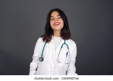 Beautiful Smiling Doctor Female With Broad Smile, Shows White Teeth, Wears Medical Uniform, Stands Over Gray Studio Wall Rejoices Having Day Off.