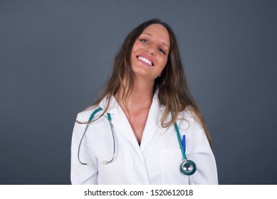 Beautiful Smiling Doctor Female With Broad Smile, Shows White Teeth, Wears Medical Uniform, Stands Over Gray Studio Wall Rejoices Having Day Off.