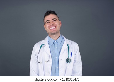 Beautiful Smiling Doctor Man With Broad Smile, Shows White Teeth, Wears Medical Uniform, Stands Over Gray Studio Wall Rejoices Having Day Off.