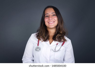 Beautiful Smiling Doctor Woman Wearing Medical Uniform With Broad Smile Stands Over Gray Studio Wall Rejoices Having Day Off. 