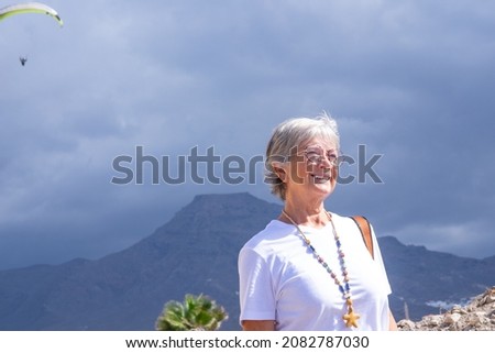 Beautiful smiling elderly woman wearing eyeglasses in outdoor excursion. A hang glider is landing behind her