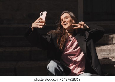 Beautiful smiling fashionable businesswoman sitting on the stairs in front of old town and make selfie, hold mobile phone, look happy. Amazed woman look happy in sunny day, show v sign, peace.