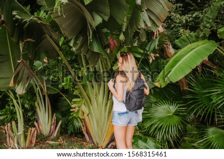 Beautiful smiling female blonde backpacker tourist  in jungle with palms. Attractive excited travel woman posing near banana tree with flower.  
