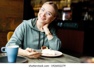 Beautiful smiling female in cafe drink coffee and eating cake. Young woman in casual clothes and glasses with smartphone in hands.