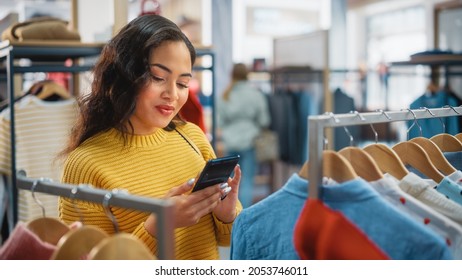 Beautiful Smiling Female Customer Shopping In Clothing Store, Using Smartphone, Browsing Online, Comparing On Internet, Choosing Stylish Clothes. Fashionable Shop, Colorful Brands, Sustainable Designs