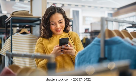 Beautiful Smiling Female Customer Shopping In Clothing Store, Using Smartphone, Browsing Online, Comparing On Internet, Choosing Stylish Clothes. Fashionable Shop, Colorful Brands, Sustainable Designs