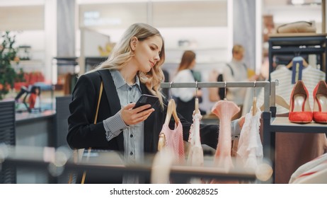 Beautiful Smiling Female Customer Shopping In Clothing Store, Using Smartphone, Browsing Online, Comparing On Internet, Choosing Stylish Clothes. Fashionable Shop, Colorful Brands, Sustainable Designs