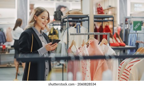 Beautiful Smiling Female Customer Shopping In Clothing Store, Using Smartphone, Browsing Online, Comparing On Internet, Choosing Stylish Clothes. Fashionable Shop, Colorful Brands, Sustainable Designs