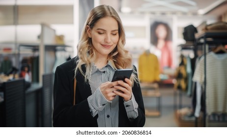 Beautiful Smiling Female Customer Shopping In Clothing Store, Using Smartphone, Browsing Online, Comparing On Internet, Choosing Stylish Clothes. Fashionable Shop, Colorful Brands, Sustainable Designs