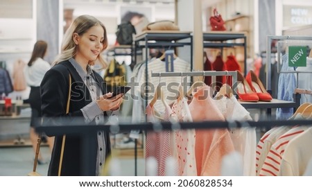 Beautiful Smiling Female Customer Shopping in Clothing Store, Using Smartphone, Browsing Online, Comparing on Internet, Choosing Stylish Clothes. Fashionable Shop, Colorful Brands, Sustainable Designs