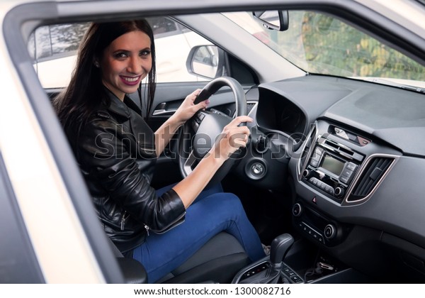 Beautiful smiling female driver in black jacket\
sitting inside car and holding steering wheel. A look into the\
vehicle window to happy attractive brunette driving automobile.\
Excited female driver