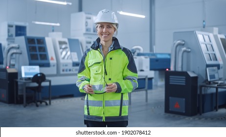 Beautiful Smiling Female Engineer Wearing Safety Vest and Hardhat Holds Safety Goggles. Professional Woman Working in the Modern Manufacturing Factory. Facility with CNC Machinery and robot arm