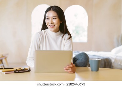 Beautiful Smiling Freelancer Asian Woman Working Remotely On Laptop And Looking Away In Home Winter Atmosphere. Positive Female Entrepreneur Working On New Project At Desk. Working From Home Concept.