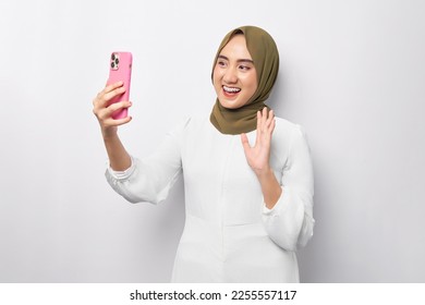 Beautiful smiling friendly Arabian Asian Muslim woman in hijab green doing video call on mobile phone, waving greeting with hand on white background studio portrait. People religious lifestyle concept - Powered by Shutterstock