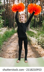 A Beautiful, Smiling Girl Athlete Professional, A Child Cheerleader In A Black Suit Stands On A Pedestal Raises His Hands Up With Orange Pom-poms, Victory. Cheerleading Champion.