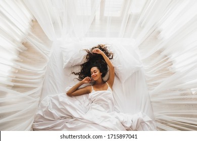 Beautiful smiling girl awakening in white bed. Happy wake up and start new day. Leisure and rest. VIew from above. Wellbeing concept.