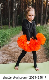 A Beautiful, Smiling Girl, A Child Cheerleader In A Black Suit Dances, Performing Movements, In The Forest On A Green Rug With Orange Pom-poms In Her Hands. Sports Training For Cheerleading.