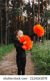 A Beautiful, Smiling Girl, A Child Cheerleader In A Black Suit Dances, Performing Movements, In The Forest On A Green Rug With Orange Pom-poms In Her Hands. Sports Training Cheerleading.