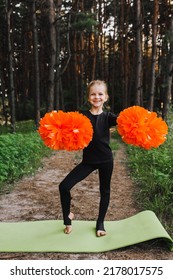 A Beautiful, Smiling Girl, A Child Cheerleader In A Black Suit Dances, Performing Movements, In The Forest On A Green Rug With Orange Pom-poms In Her Hands. Sports Training Cheerleading.