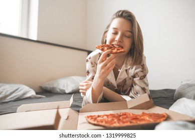 Beautiful smiling girl in a pajama lies in the bed of a home with a box of pizza, eating a piece of pizza with her eyes closed. Happy girl eating pizza for breakfast in bed. Fast food. Copyspace