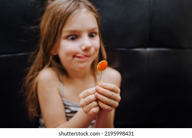 A Beautiful, Smiling Girl Of Preschool Age, A Red-haired Child, A Sweet Tooth, Sits On A Black Leather Sofa With A Lollipop In Her Hands And Looks At Him, Dreaming Of Eating. Happy Childhood.