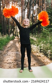 A Beautiful, Smiling Girl, A Professional Athlete, A Child Cheerleader In A Black Suit Stands On A Pedestal Raises His Hands Up With Orange Pom-poms, Delighted With The Victory. Cheerleading Champion.